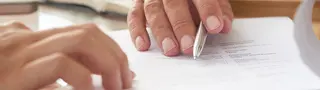Eine Person hat die Hand auf einem Tisch abgelegt. In der Hand wird ein Stift gehalten.